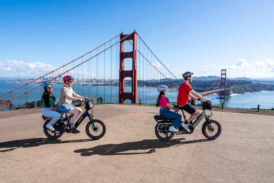 Two riders smiling with electric bikes in front of the bridge