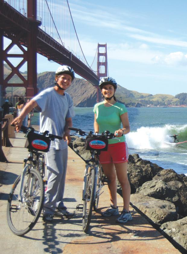 Couple posing with their bikes underneath the Golden Gate Bridge with surfers on the water